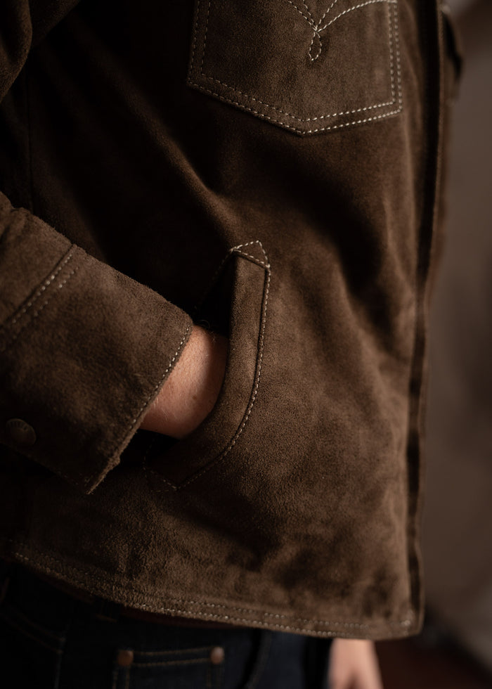 A person wearing a brown suede jacket with detailed stitching, hand casually tucked into the pocket, creating a relaxed look.