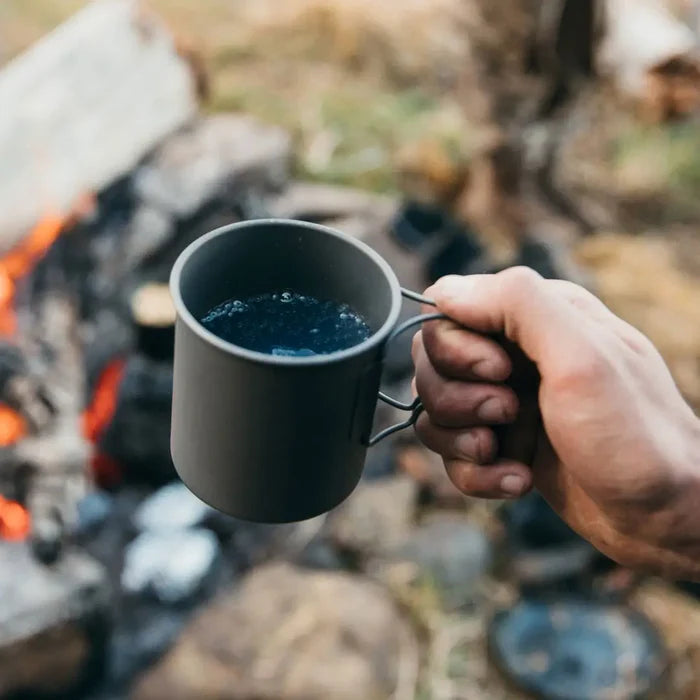 Hand holding a Lifeventure Titanium Camping Mug by the handles with a blurred campfire background