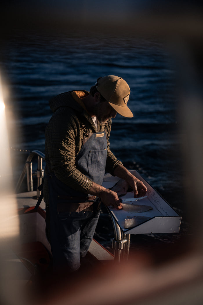 A person in a cap and overalls fillets fish on a boat at sunset, with dark ocean waves in the background.