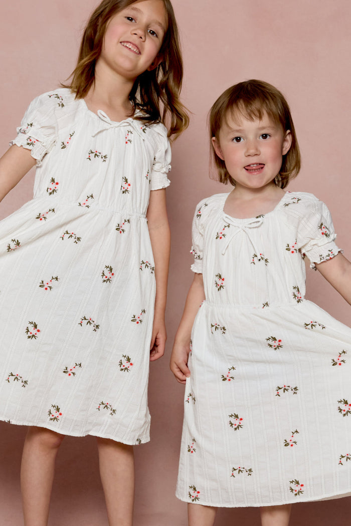 Two young children stand side by side, smiling in front of a pink background while wearing the Ivy City Co Mini Jessie Dress in Ditsy Embroidery.