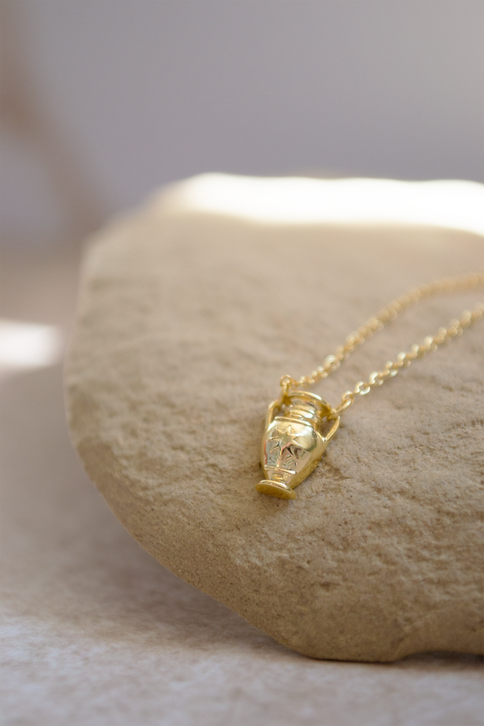 A gold necklace with a small sculpted pendant resting on a smooth beige stone against a soft neutral background.
