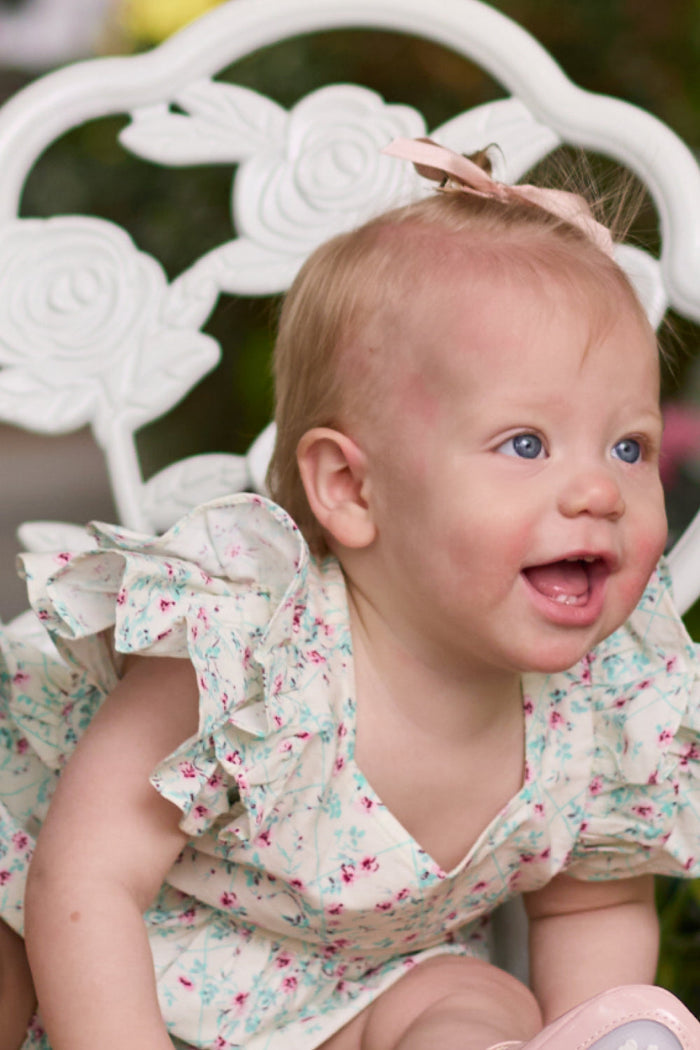 A smiling baby with blue eyes and light hair sits on a white floral-patterned chair, wearing the ruffled Baby Hattie Romper in Pansy Trellis by Ivy City Co and a small pink bow in her hair.