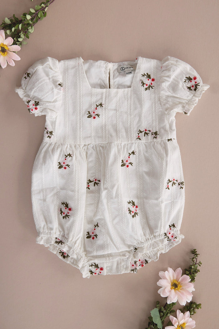 The Ivy City Co Baby Jessie Romper in Ditsy Embroidery is a white cotton romper with puffed sleeves, pintuck details, and pink and green floral accents. Shown flat on a beige background with daisies and greenery.