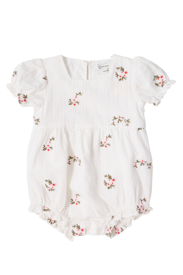 The Ivy City Co Baby Jessie Romper in Ditsy Embroidery is a white cotton romper with pink and green floral embroidery, short puffed sleeves, textured stripes, ruffled legs, gathered waist, smocked bodice, and back buttons.
