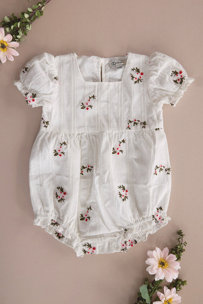 The Baby Jessie Romper in Ditsy Embroidery by Ivy City Co, featuring a smocked bodice, puff sleeves, delicate floral embroidery, and elastic leg openings, is displayed on a beige surface adorned with pink and white flowers and green foliage around the edges.