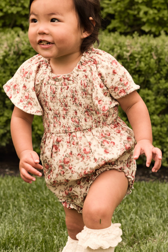 A young child in the Ivy City Co Baby Jovie Romper in Ivory Heritage Rose and cream socks plays on green grass with bushes behind, smiling—perfect for adorable Mom and Me matching moments.
