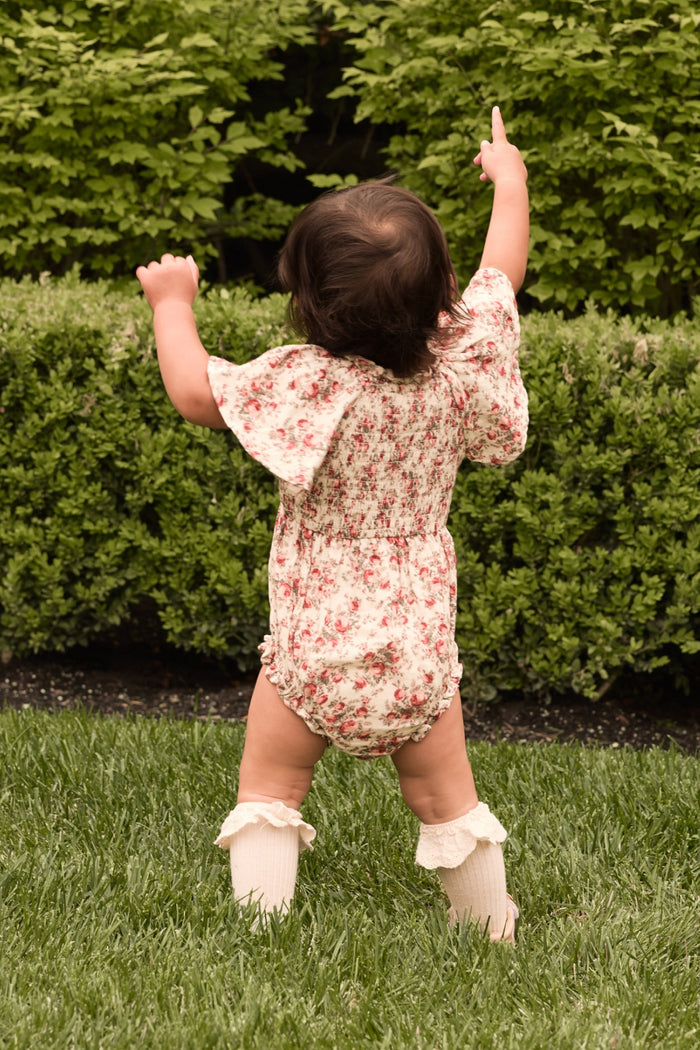 A baby girl wearing the Ivy City Co Baby Jovie Romper in Ivory Heritage Rose and cream socks stands on grass, facing away and pointing with both hands toward green bushes.