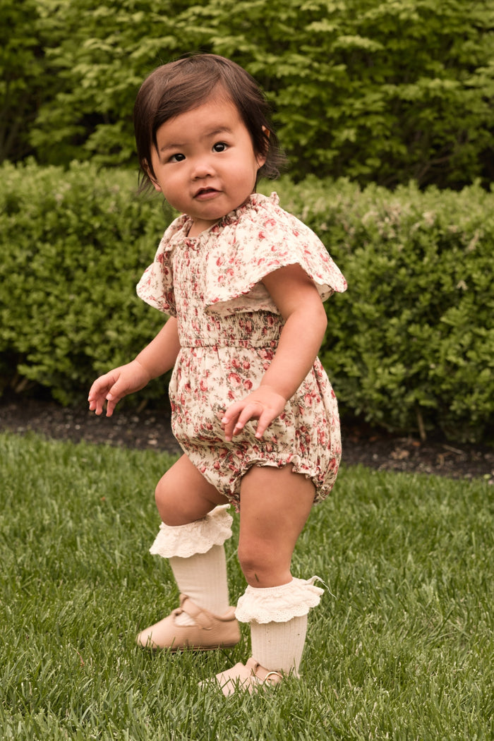 A toddler wears the Ivy City Co Baby Jovie Romper in Ivory Heritage Rose with cream knee-high socks and beige shoes, standing on green grass surrounded by lush bushes.