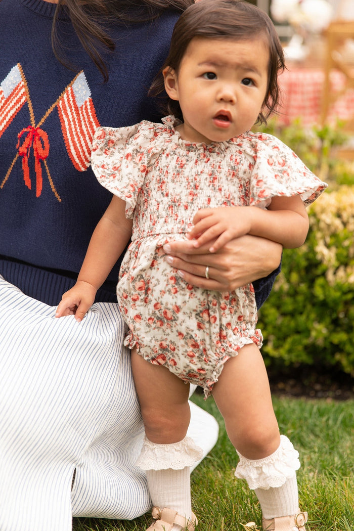 A toddler wearing Ivy City Co’s Baby Jovie Romper in Ivory Heritage Rose, featuring ruffled sleeves, stands on the grass with support from an adult. The adult wears a navy sweater with flag designs and a white skirt, creating a subtle Mom and Me matching moment against a backdrop of greenery and outdoor seating.