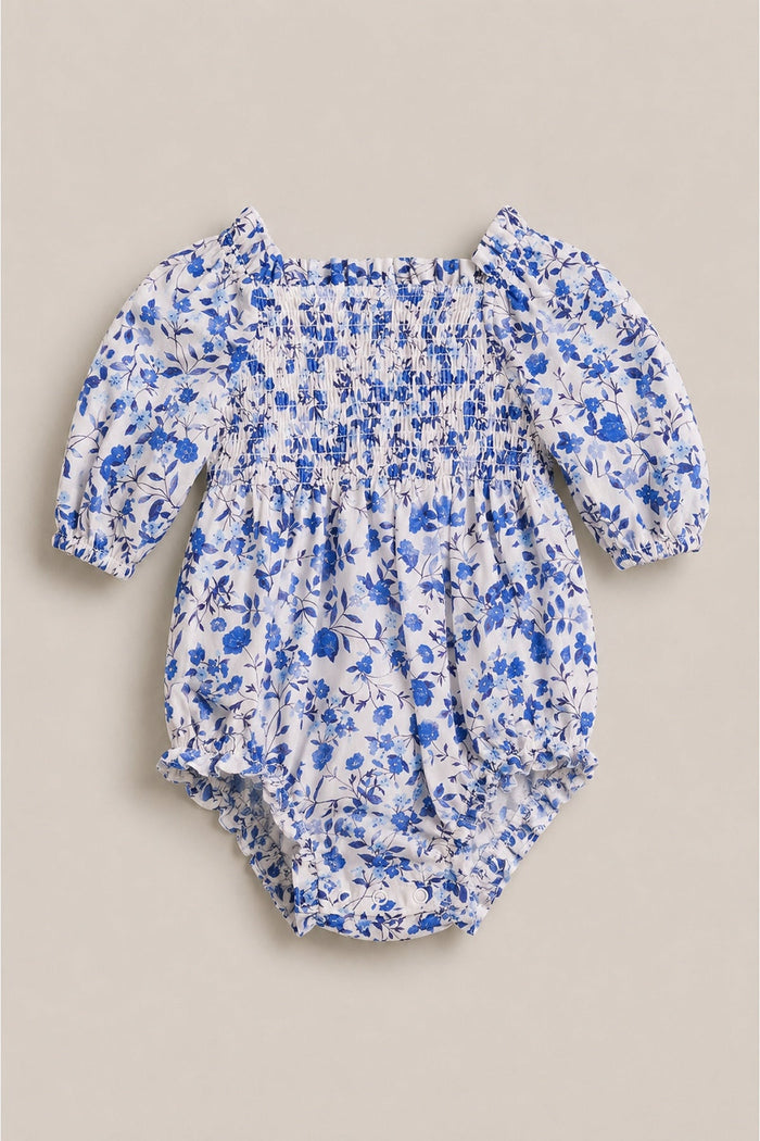 The Ivy City Co Baby Madeline Romper in Ditsy Cobalt Floral features a blue and white floral print, smocked bodice, puffy long sleeves, and elastic leg openings, displayed on a neutral background.