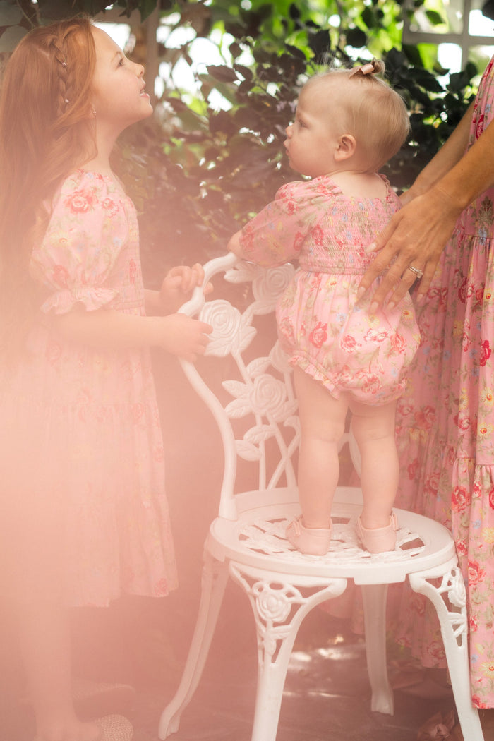 A young girl and a toddler in matching pink floral outfits interact by a white ornate chair, while an adult’s hand gently supports the toddler, who is wearing the Ivy City Co Baby Madeline Romper in Pink Vintage Poppies with a smocked bodice. Soft, dreamy lighting fills the scene.