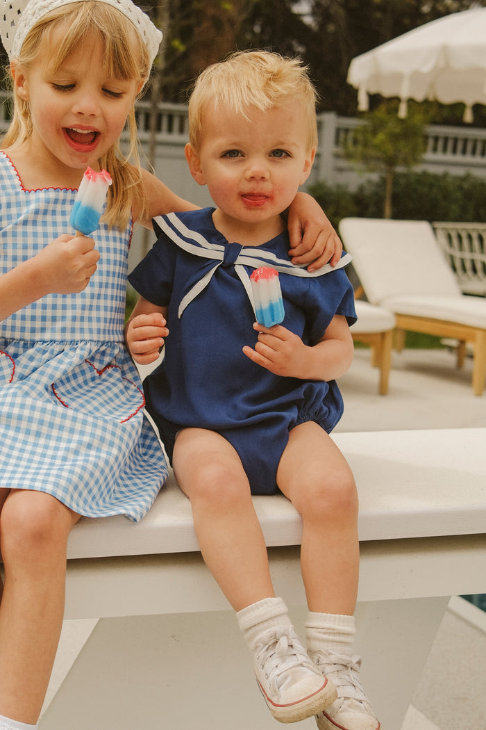 Two young children sit by a pool, smiling and eating red, white, and blue popsicles. One wears a blue gingham dress; the other is in the Ivy City Co Baby Sailor Romper in Navy with white sneakers for the perfect nautical look.