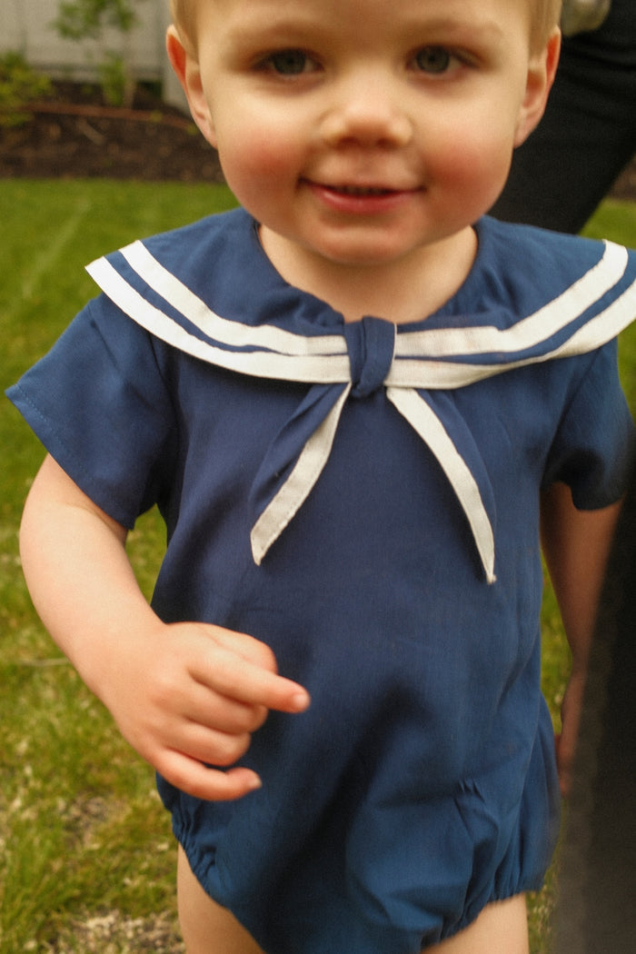 A smiling toddler wears the Ivy City Co Baby Sailor Romper in Navy, featuring white trim, while standing on grass. An adult's arm is visible in the background.