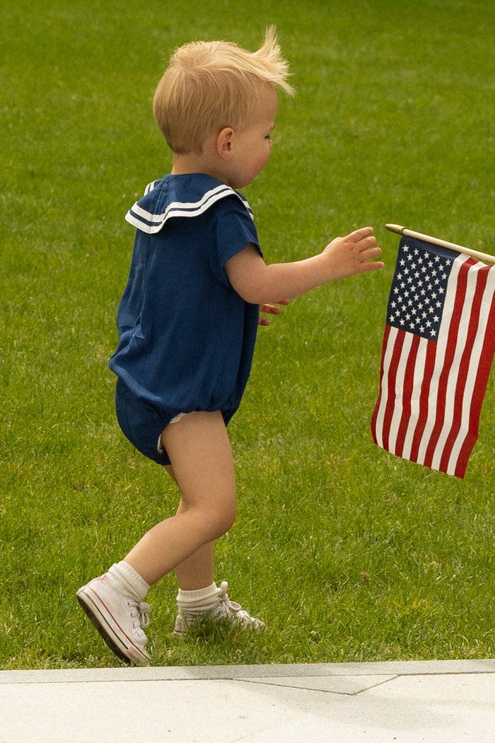 A young child in the Ivy City Co Baby Sailor Romper in Navy and white sneakers walks on grass toward a small American flag on a stick.