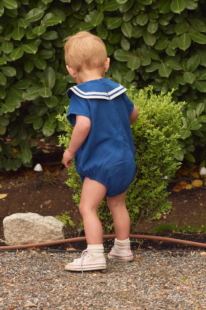 A small child wears the Ivy City Co Baby Sailor Romper in Navy with white shoes, standing outdoors on gravel near a green bush and leafy plants, facing away from the camera.