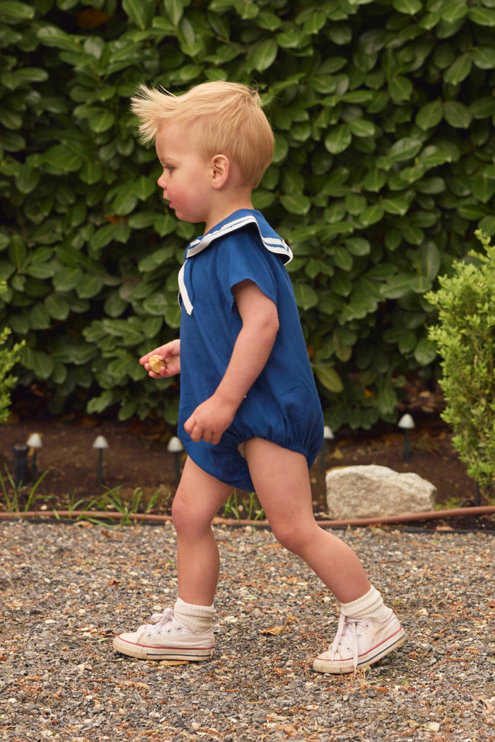 A young child with blonde hair walks outside on a gravel path, wearing the Ivy City Co Baby Sailor Romper in Navy with a sailor collar, white socks, and white sneakers. Green bushes and small plants are visible in the background.