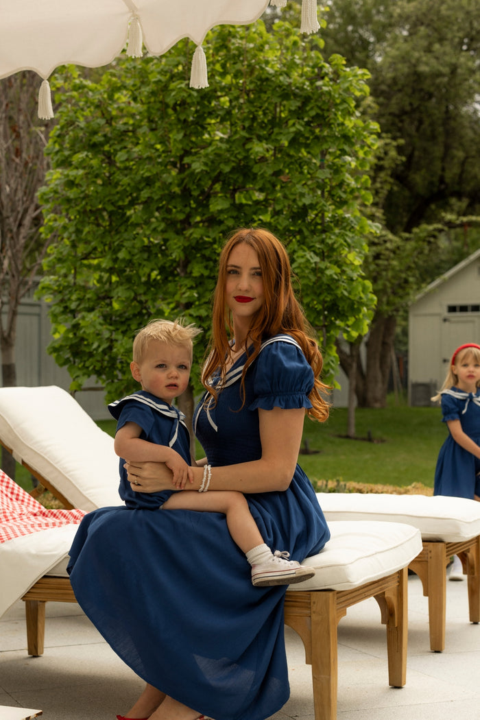 A woman in a blue dress sits outdoors, holding a young child wearing the Ivy City Co Baby Sailor Romper in Navy. A girl in a matching blue dress stands near greenery in the background.