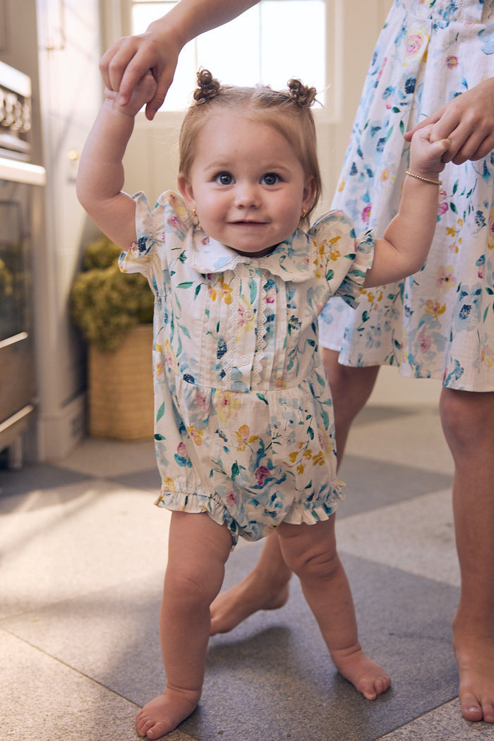 A toddler wearing the Ivy City Co Baby Sophia Romper in Blue Bouquet smiles while holding an adult's hands for support, as another person in a floral dress stands nearby with sunlight streaming through the window behind them.