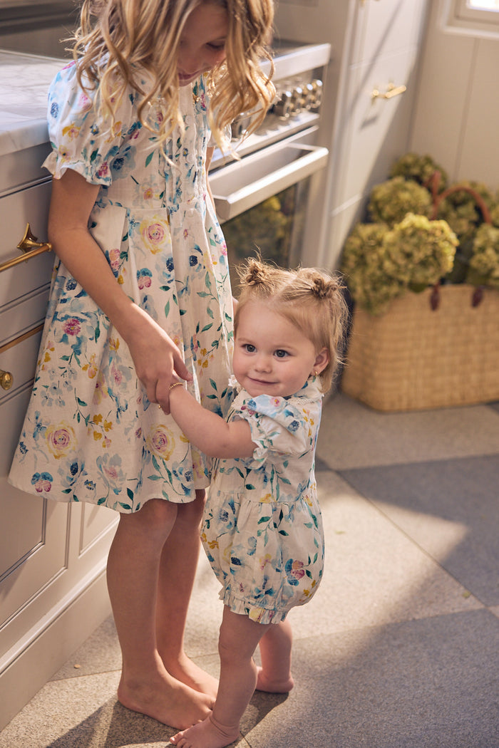 A toddler wears the Ivy City Co Baby Sophia Romper in Blue Bouquet, holding hands with an older girl in a matching floral dress. Both are barefoot in a sunlit kitchen, with a basket adorned by a blue bouquet completing the charming scene.