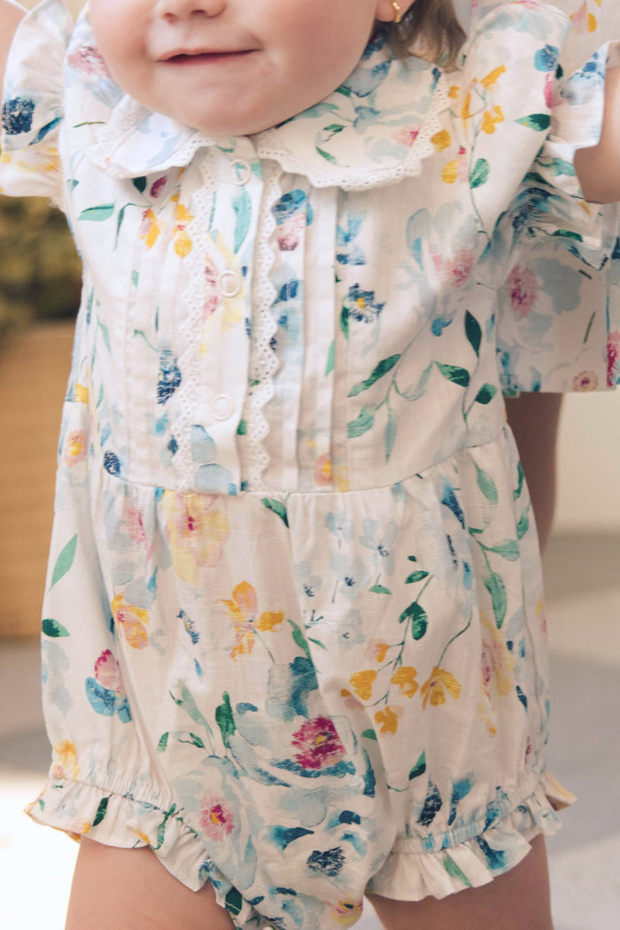 A young child wears the Ivy City Co Baby Sophia Romper in Blue Bouquet, featuring ruffled sleeves, a ruffled collar, white front buttons, and a charming blue floral print. The smiling child's face is partly out of frame.