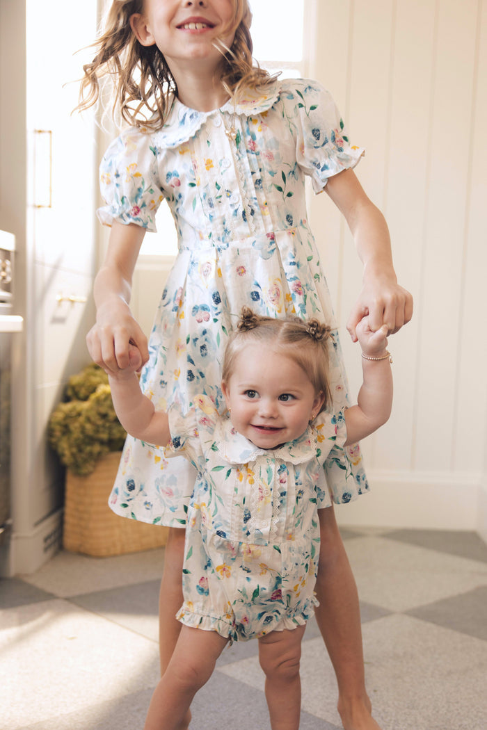 Two young girls smile and hold hands in a sunlit room. The toddler wears the Ivy City Co Baby Sophia Romper in Blue Bouquet, featuring a light floral print and ruffled collar, with a basket and light walls in the background.