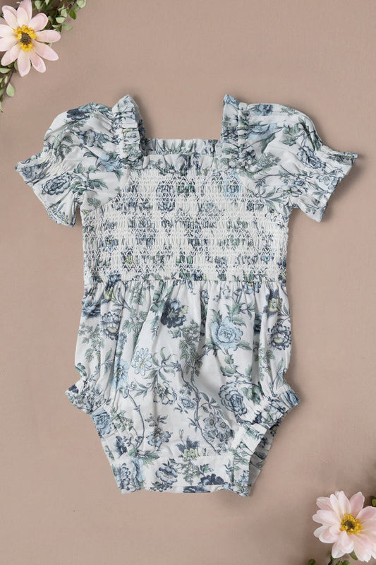 The Ivy City Co Baby Valencia Romper in Ivory Climbing Vine Bloom, made from 100% cotton with ruffled sleeves and a smocked bodice, features a blue and green floral print on ivory. Shown laid flat on beige with pink flowers nearby.