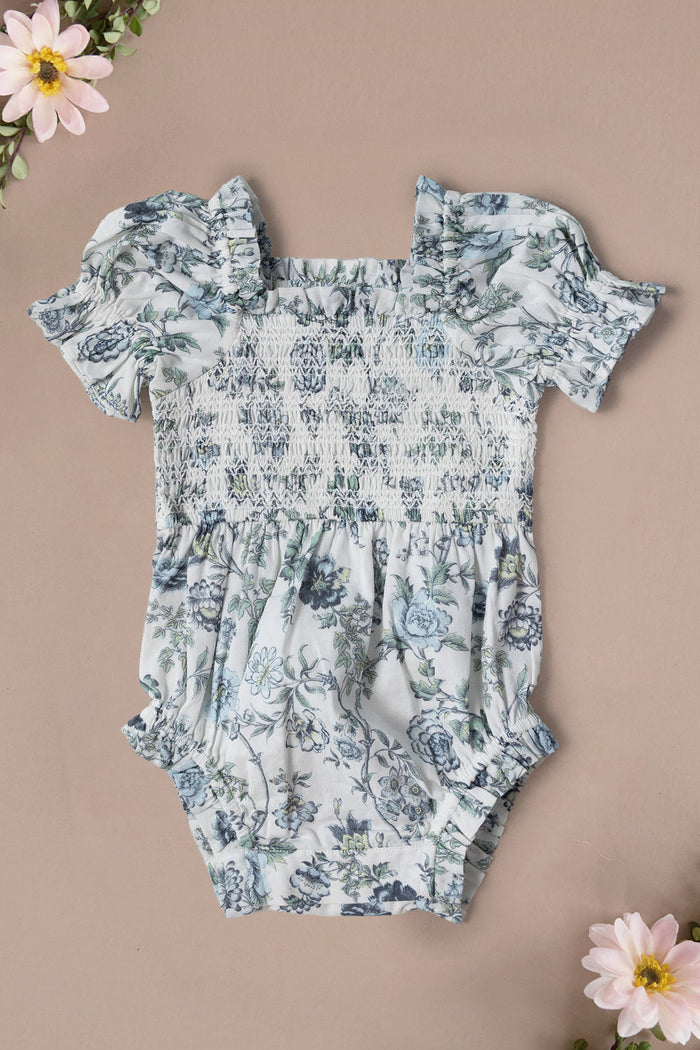 Ivy City Co’s Baby Valencia Romper in Ivory Climbing Vine Bloom features ruffled sleeves, a smocked bodice, and a gathered waist, shown on a beige background accented with pink flowers in the corners.