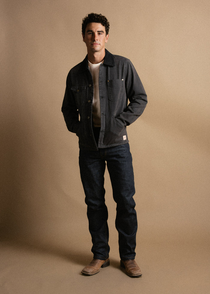 Man in denim jacket and jeans stands confidently against a beige background, hands in pockets, wearing brown boots.