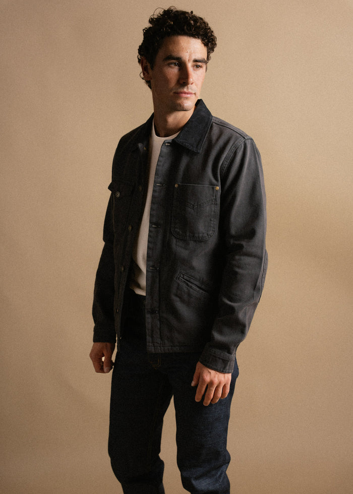 Man in a dark denim jacket and jeans stands against a beige background, looking to the side with a neutral expression.