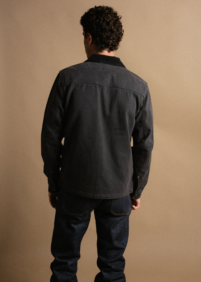 Man in dark jacket and jeans stands with back to camera against a beige background, highlighting casual fashion style.