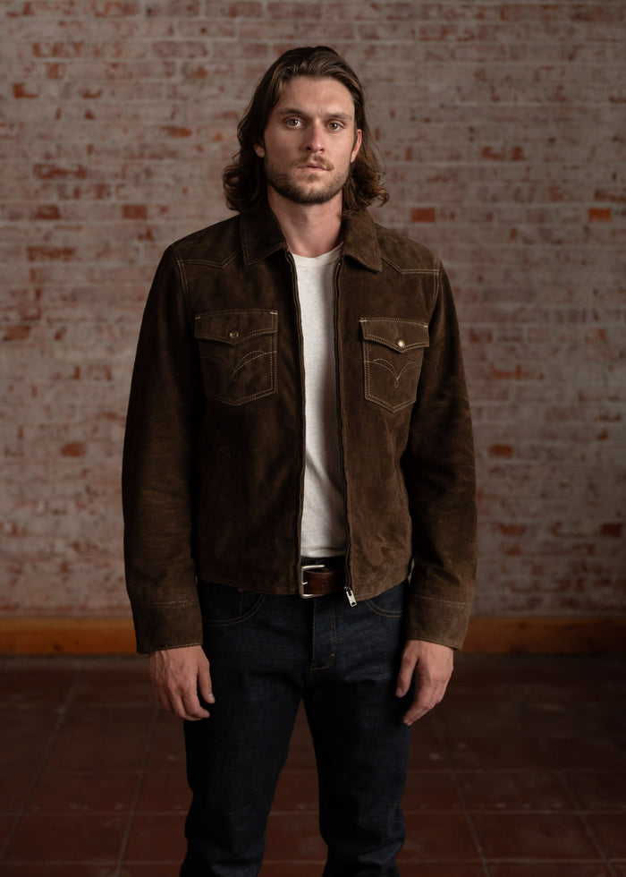 Man in a brown suede jacket and jeans stands against a rustic brick wall, exuding a casual yet confident style.