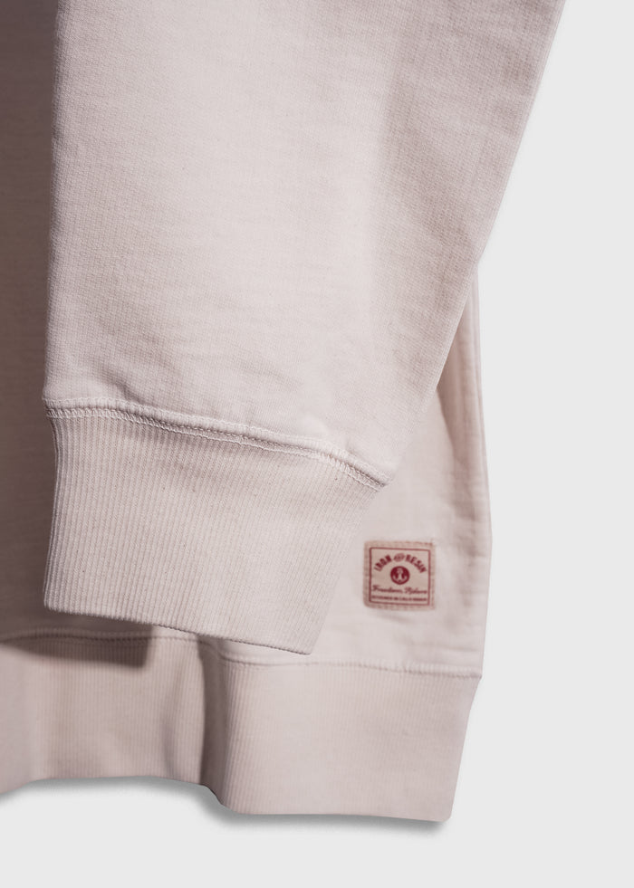 Close-up of a beige sweatshirt sleeve with ribbed cuff and a small red label near the hem on a light background.