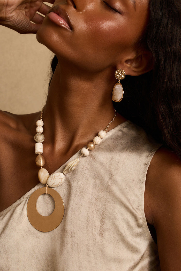 Gold beaded statement necklace with a large open-circle pendant styled over a one-shoulder top.