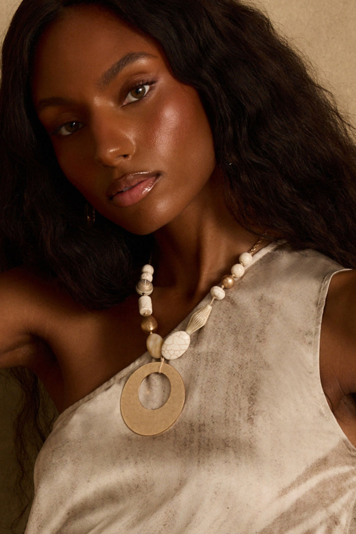 Beaded statement necklace with gold pendant and stone beads, styled with a one-shoulder top.