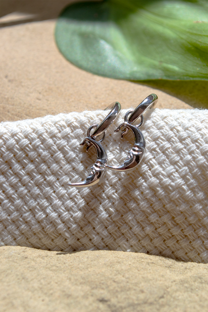 A pair of polished silver crescent moon hoop earrings resting on a textured white woven fabric, with a green leaf and sandy beige background.