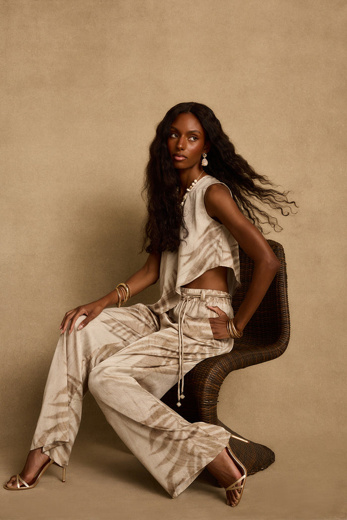 Seated on a wicker chair, a model wears the Eden palm print pant and matching top.