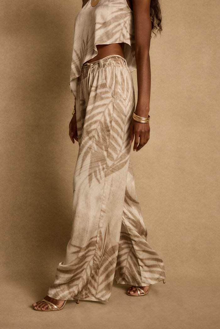 Side view of Eden palm print wide-leg satin pants with gold heels and bangles.