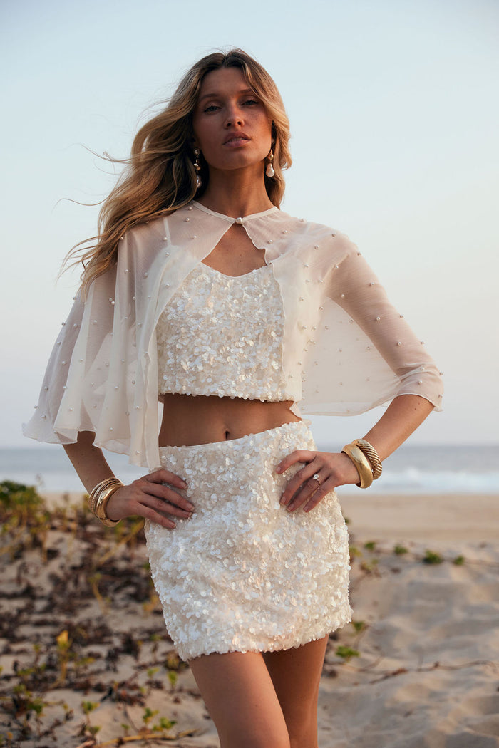 White sequin crop top and matching mini skirt styled with a pearl cape on the beach.