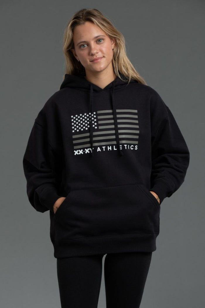Person wearing a black hoodie with a flag design and text on a gray background || Black