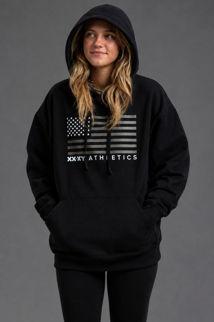 A woman wearing a black pullover hoodie with a distressed American flag graphic and 'XXXY ATHLETICS' text on the chest, posed against a solid dark gray studio background. || Black