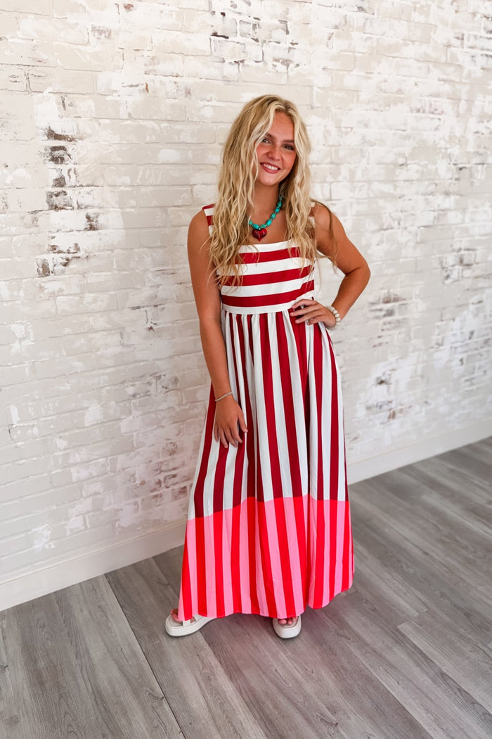 Woman wearing a sleeveless red and ivory striped maxi dress with a square neckline, flowing skirt, side pockets, and a vibrant pink color-block hem.