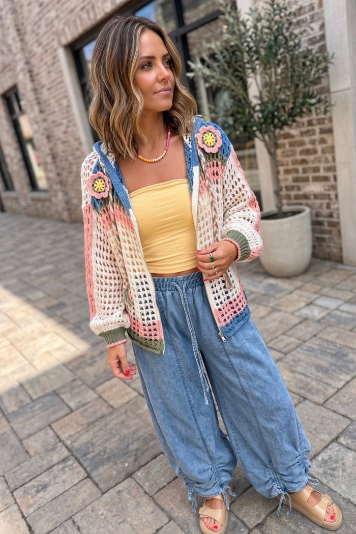 Open-knit crochet zip hoodie with colorful pink, blue, and olive stripe details, floral shoulder appliqués, and a hooded silhouette styled over a yellow tube top and chambray drawstring pants.