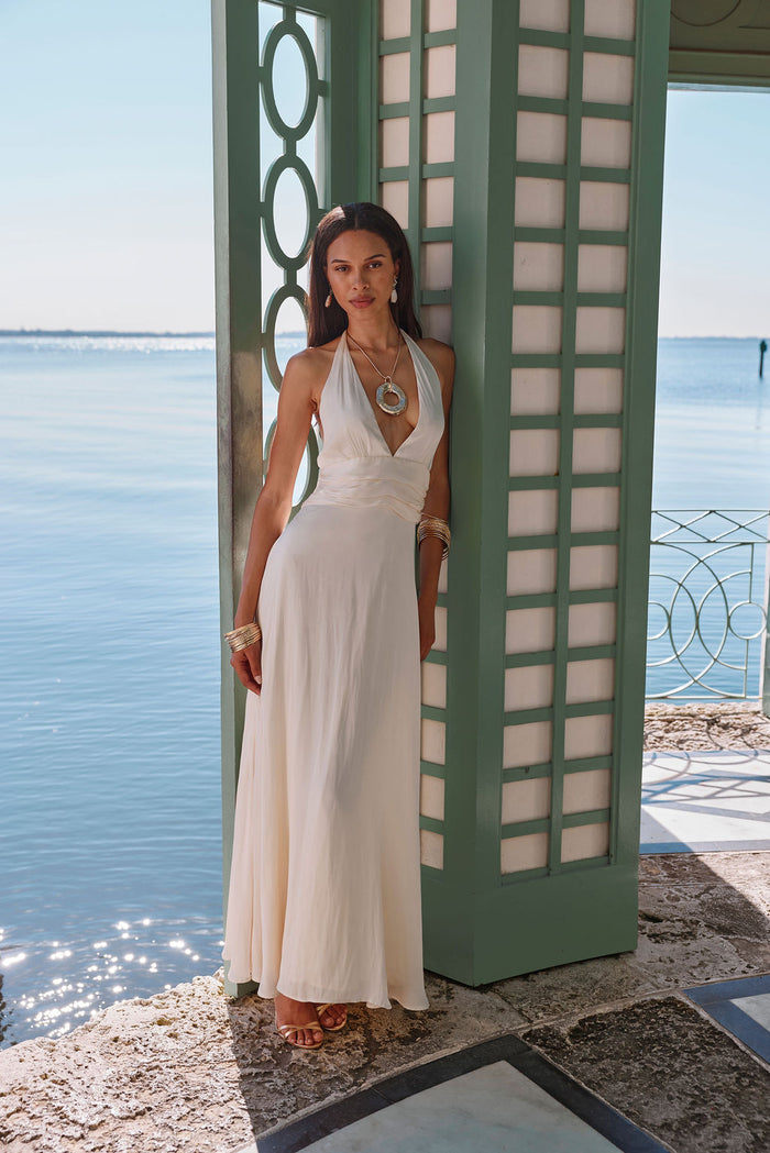 Cream satin halter maxi dress worn beside a green pavilion overlooking calm ocean water.