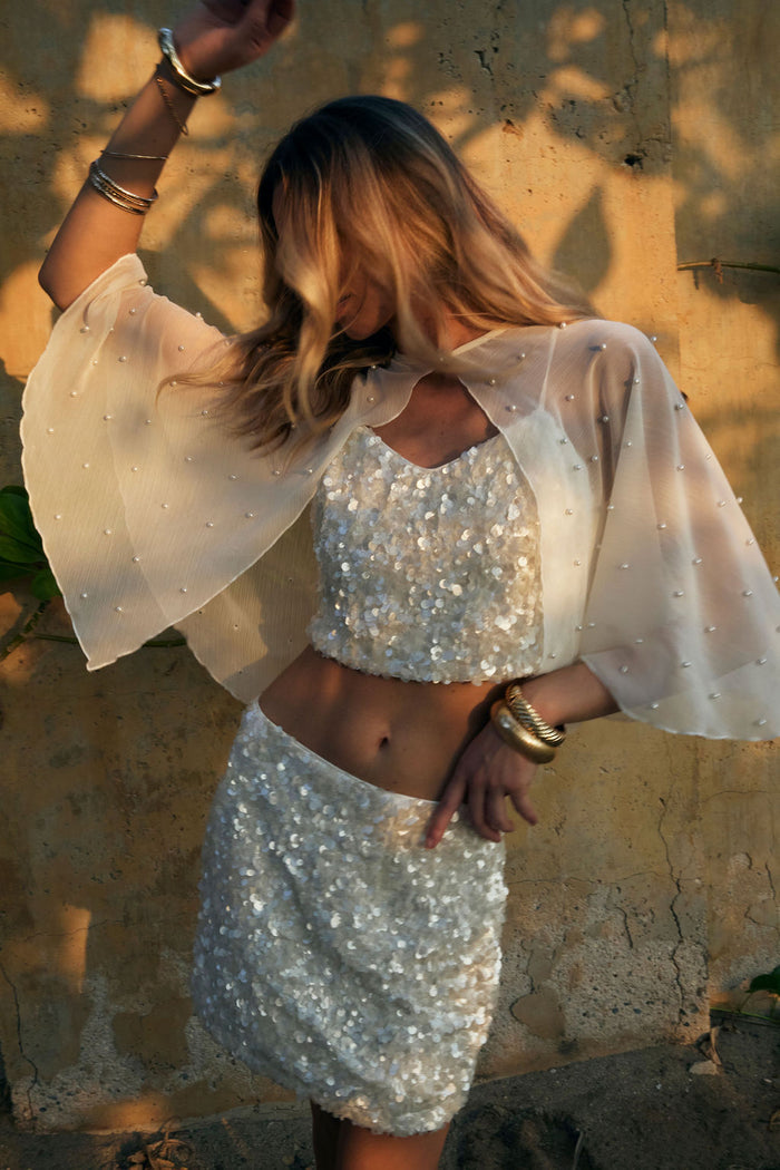 Sheer pearl-dotted white cape layered over a sequin crop top and mini skirt at golden hour.