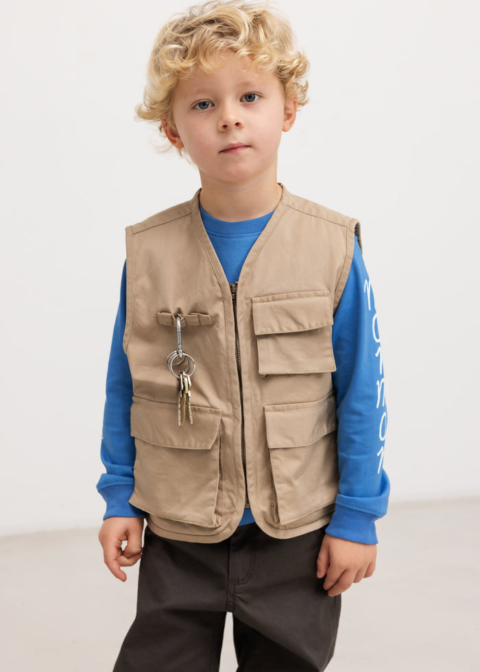 Sand-colored children's vest with snap pockets and zipper, worn over a blue blouse