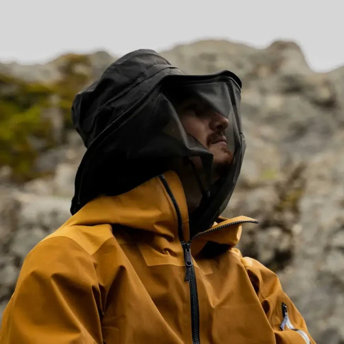 A man wearing a Lifesystems Midge And Mosquito Boonie Head Net Hat with the mesh attached, standing outdoors against a blurred rocky background.