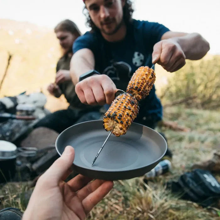 Person holding a skewer with grilled corn over a Lifeventure titanium camping plate outdoors