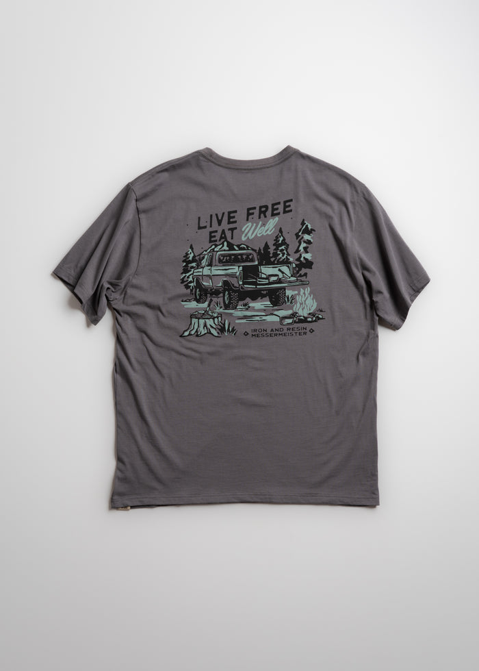 Gray T-shirt with "Live Free, Eat Well" slogan and truck graphic in forest setting. Casual outdoor adventure theme.