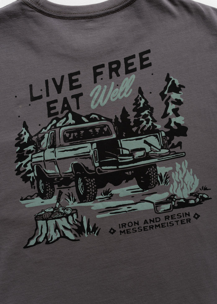 T-shirt design with a truck, trees, and campfire, featuring "Live Free Eat Well" and "Iron and Resin Messermeister" text.