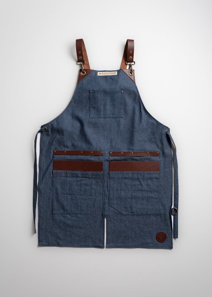 Denim apron with leather straps and pockets, featuring brass rivets, displayed against a plain background for a rustic look.